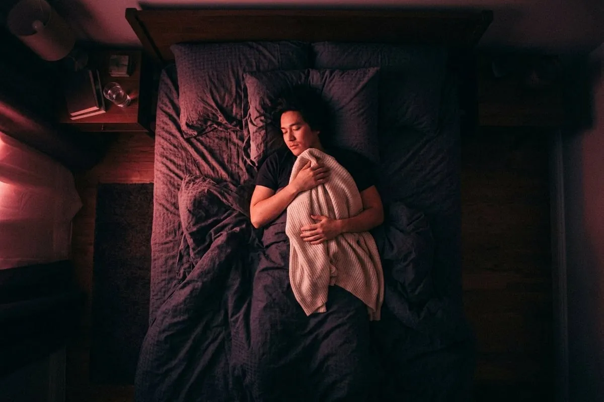 一個人獨自躺在床上，懷抱著衣物，暗室中透著一縷玫瑰色微光，情緒安靜而深沉